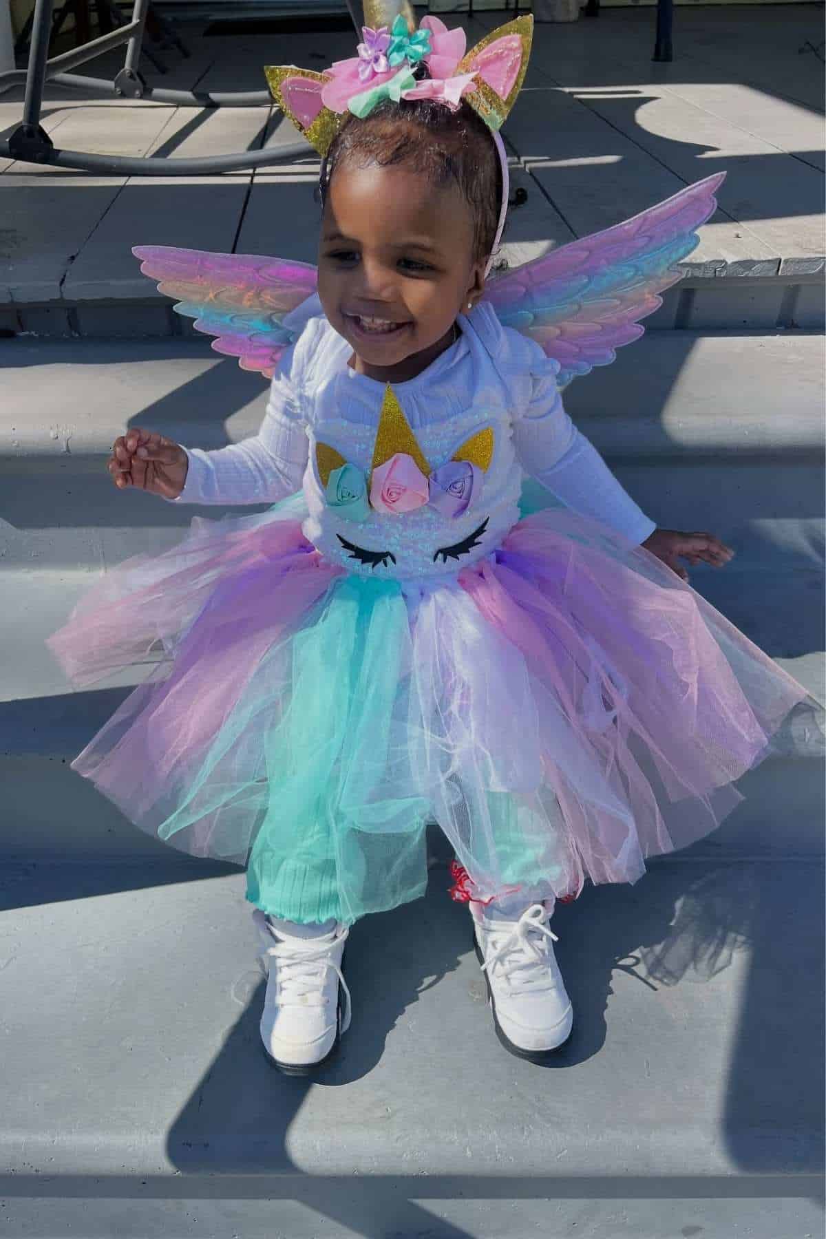 A young child dressed in a unicorn-themed costume with pastel wings, a tutu, and matching headband sits and smiles on outdoor steps.