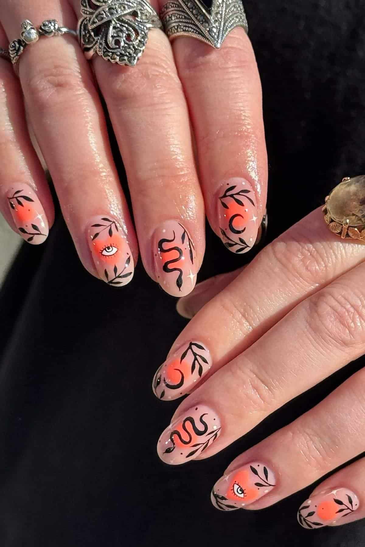 Hands with almond-shaped nails painted with orange ombre tips, black leaf designs, and black-red art of snakes, eyes, and crescent moons. Fingers have silver and gold rings.
