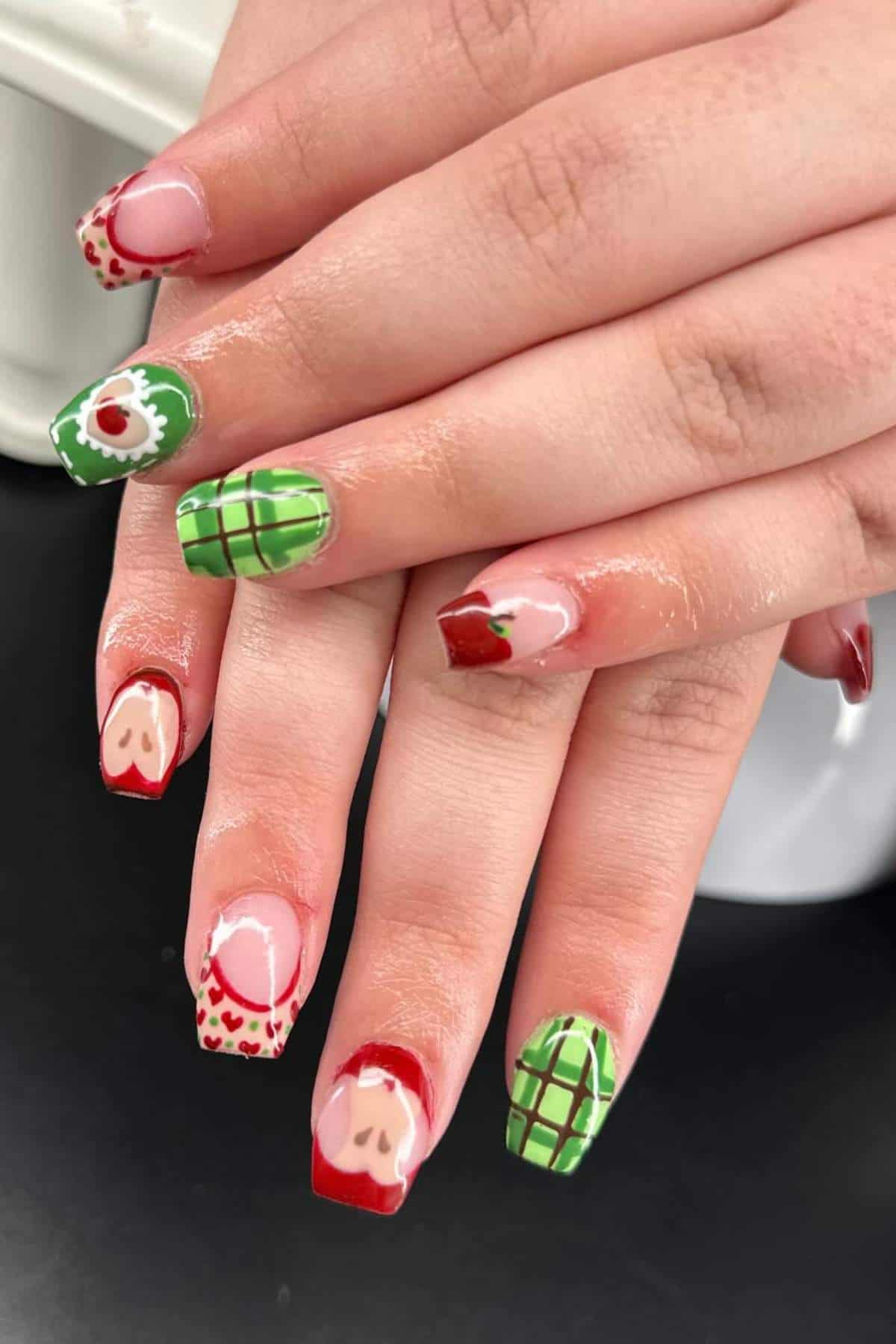 Hands with long square nails featuring various strawberry-themed nail art designs in red, green, and white, including plaid patterns, hearts, and strawberries.