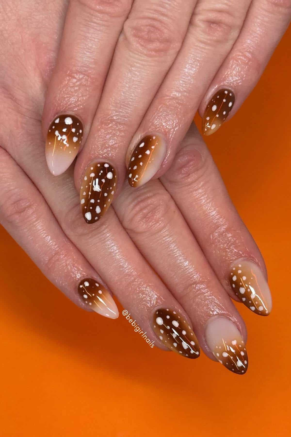 Hands with almond-shaped nails featuring brown ombre polish and white dot designs, resembling a deer pattern, against an orange background.
