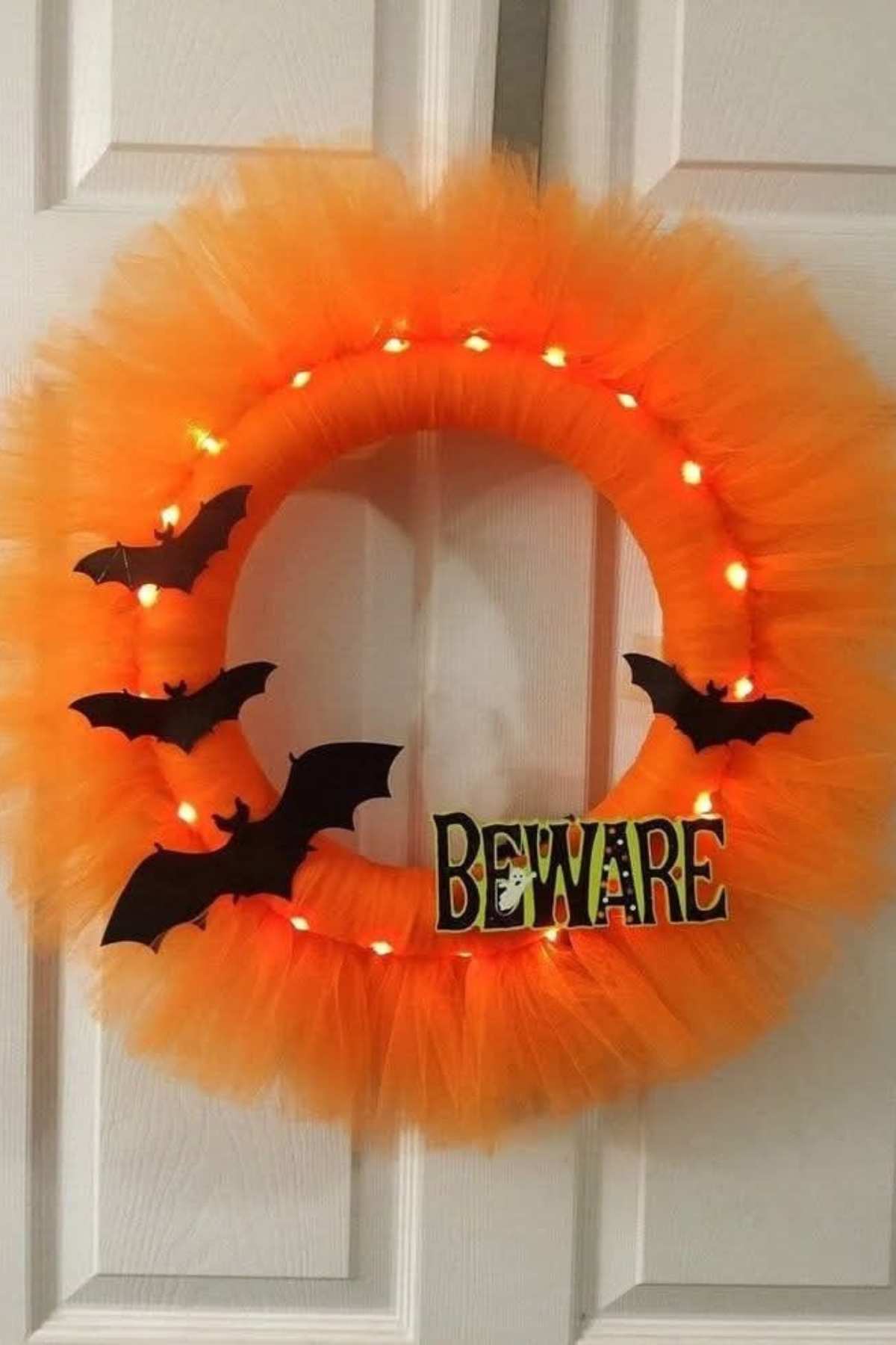 An orange tulle wreath with black bat cutouts, string lights, and a sign that reads "BEWARE," hanging on white double doors.