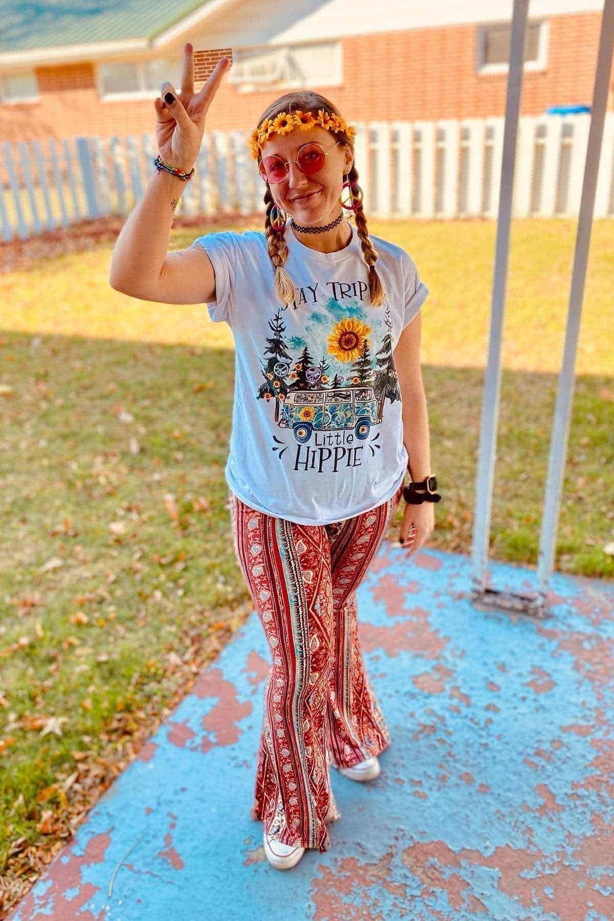 Person wearing a "Day Tripper Little Hippie" shirt, patterned bell-bottom pants, round sunglasses, flower headband, and showing a peace sign, standing outdoors on a porch.