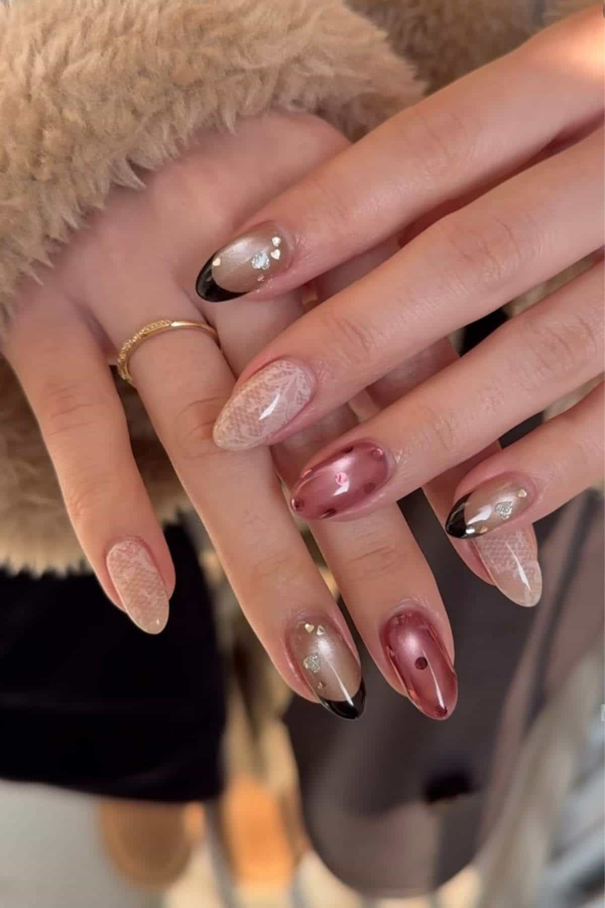 Hands with long almond-shaped nails, featuring a mix of pink chrome, lace designs, black French tips, and small embellishments.