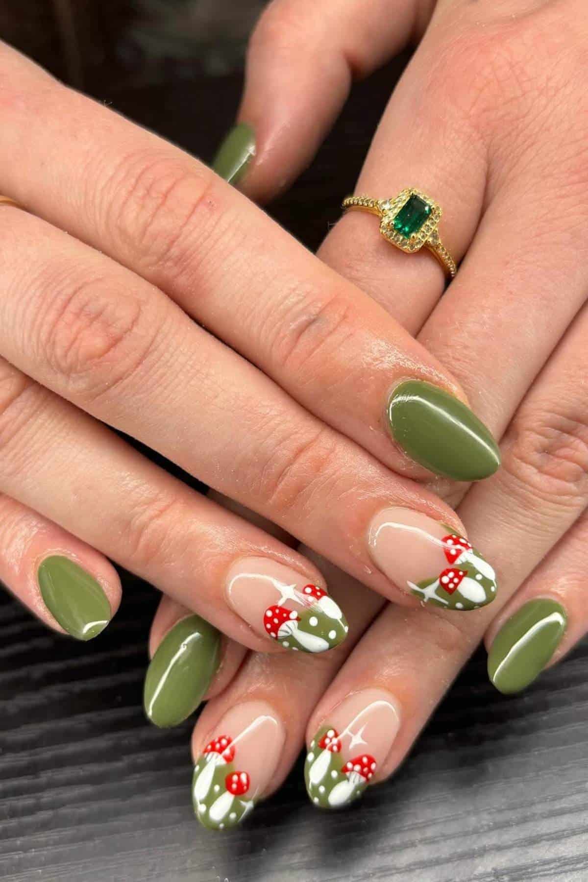 Hands with olive green manicure; some nails feature mushroom and foliage nail art. A gold ring with a green gemstone is worn on one finger.