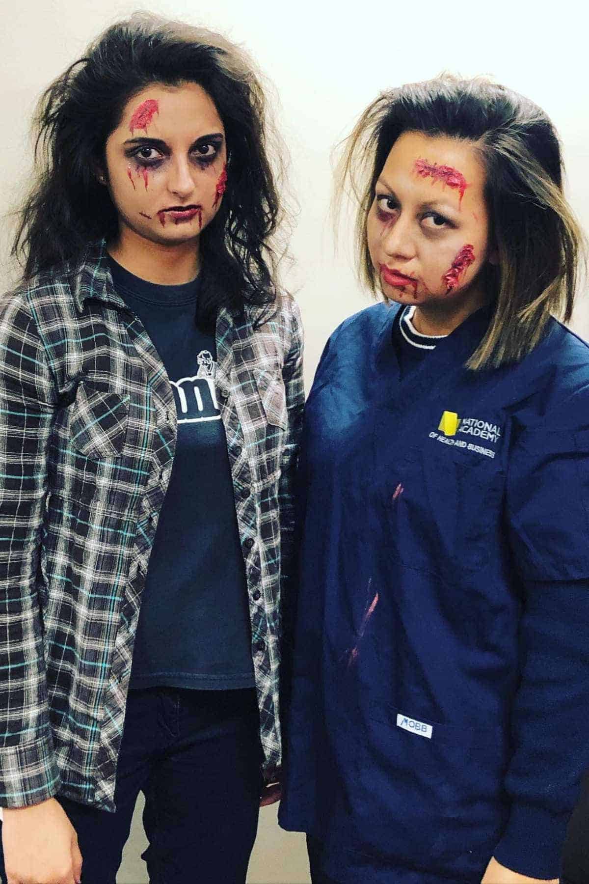 Two people wearing zombie makeup with fake wounds and blood stand side by side; one is in a plaid shirt and the other wears medical scrubs.