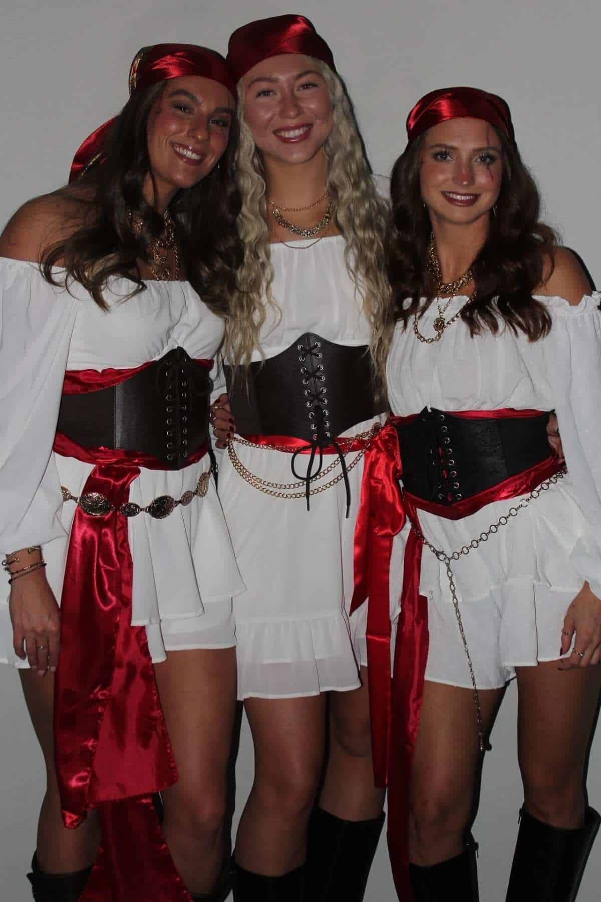 Three women stand together wearing matching pirate costumes with white dresses, red sashes, red headscarves, black belts, and black boots, posing against a plain background.