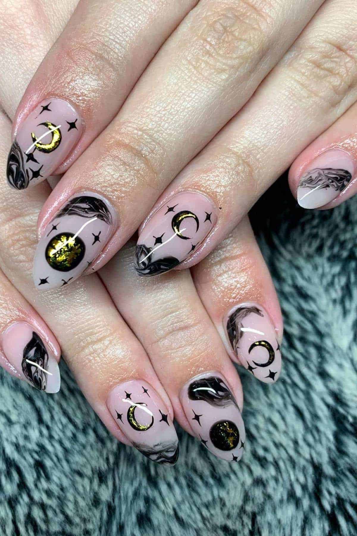 Hands with almond-shaped nails featuring black marble tips, gold crescent moons, and black star designs on a pale pink base, resting on a textured gray surface.