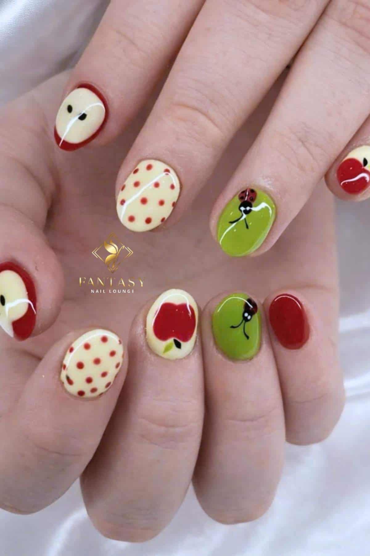 Fingernails painted with red, green, and cream polish featuring apple, polka dot, and cherry designs on a white satin background.