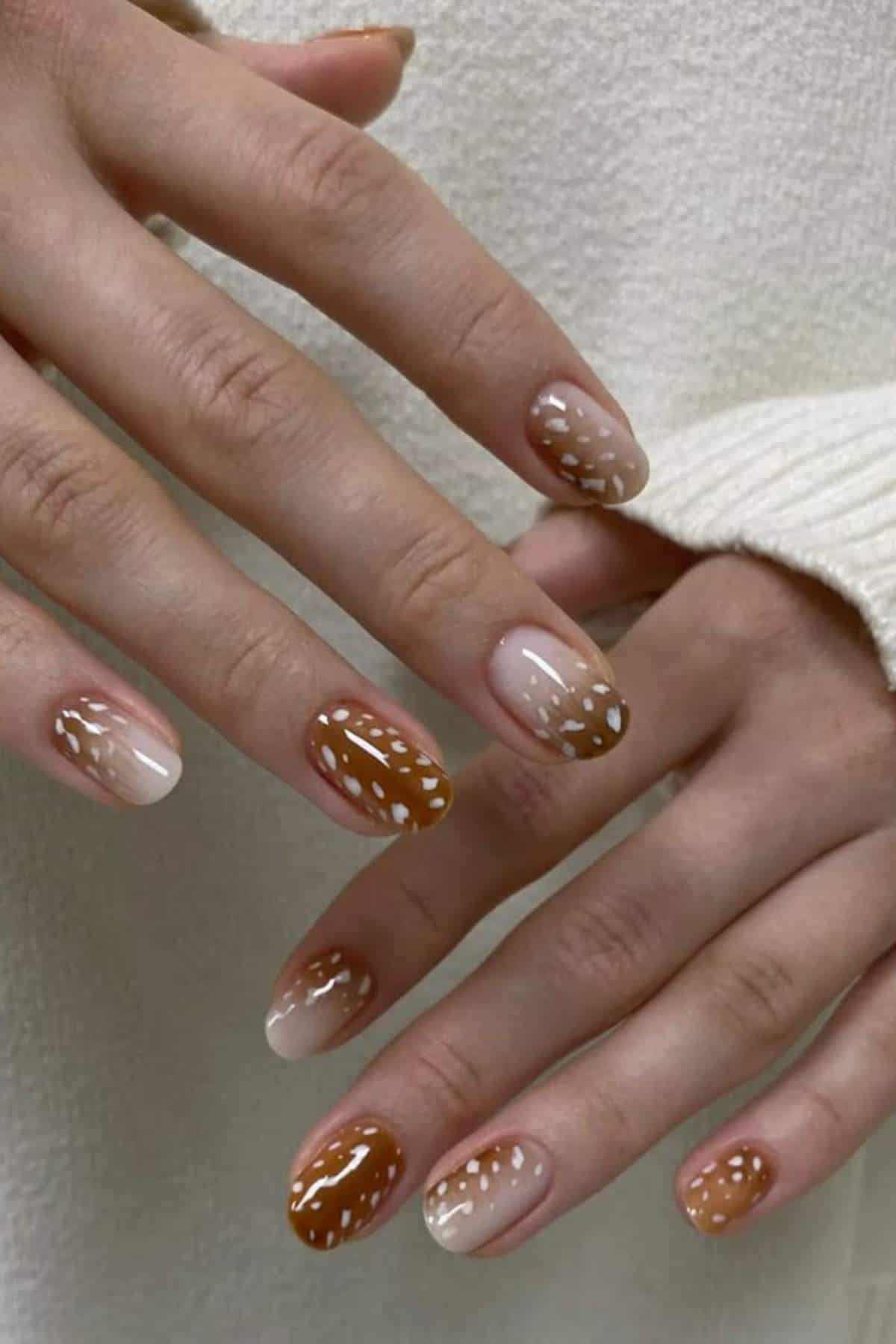 Hands with almond-shaped nails painted in brown and beige tones, featuring a white dotted pattern, against a cream-colored background.