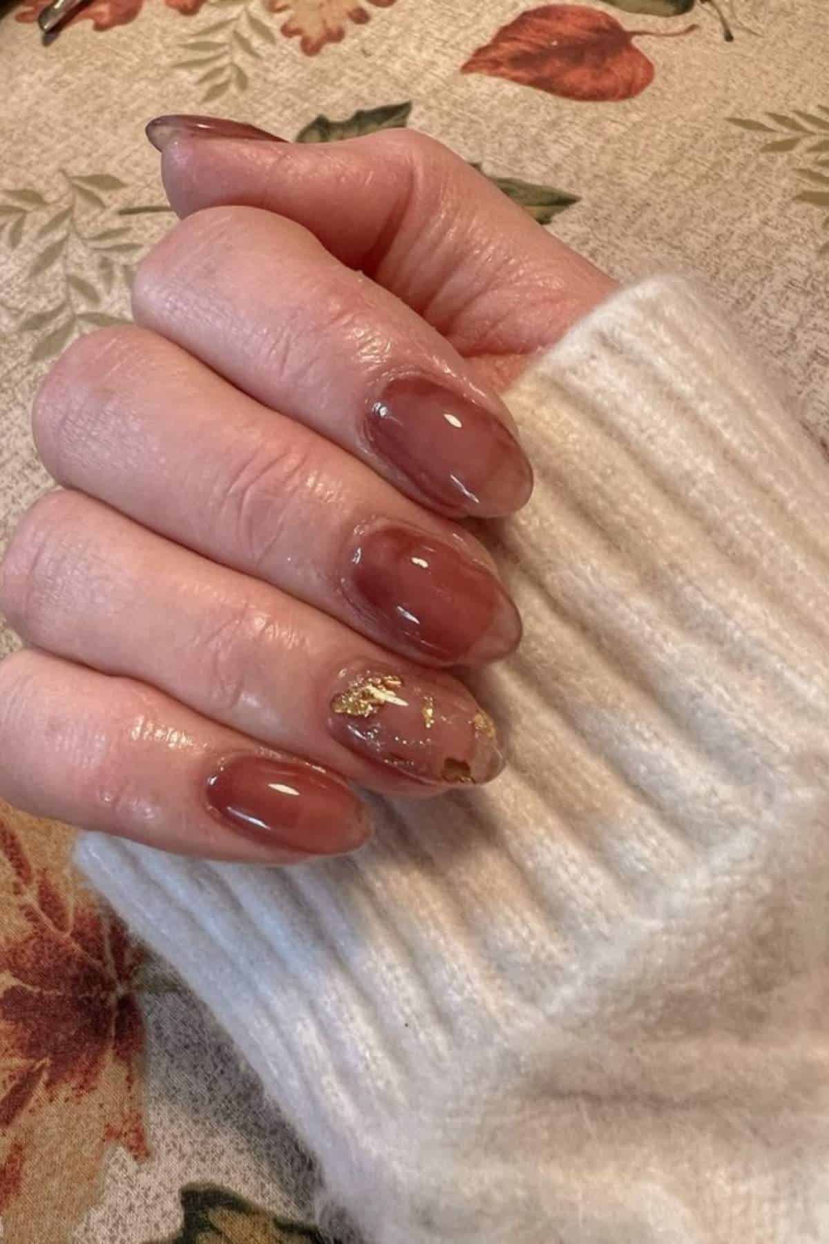 A hand with brown oval-shaped gel nails, one nail featuring gold foil accents, rests on a cream ribbed sweater sleeve over a floral-patterned surface.