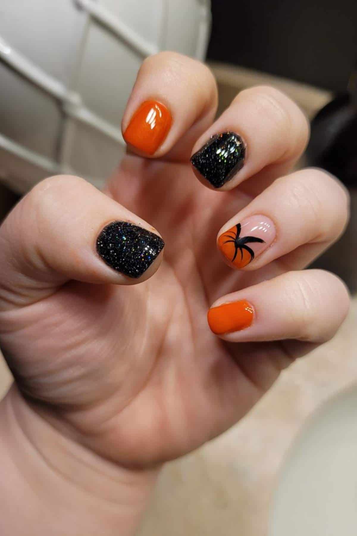 A hand with orange and glittery black nail polish; one nail features a black spider design on a nude background.