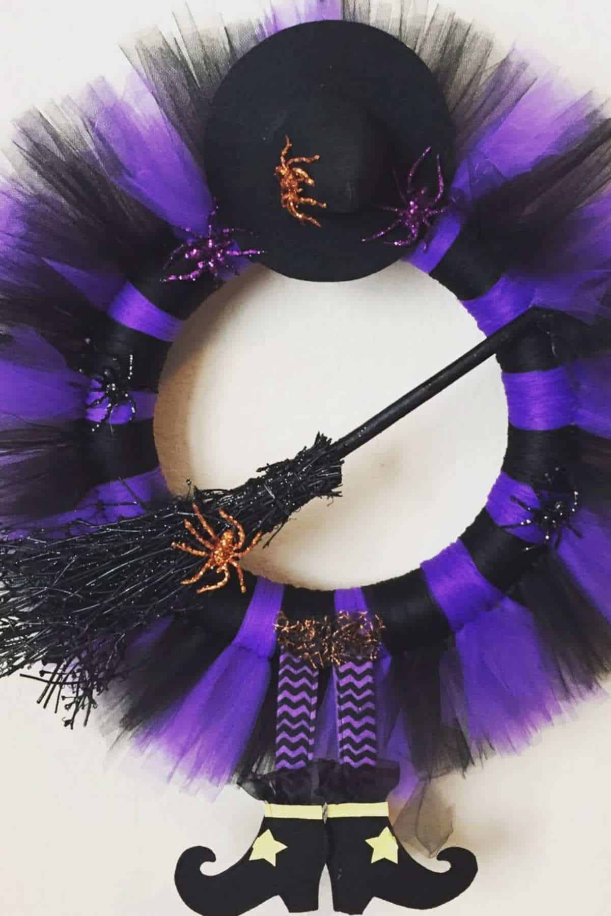 A wreath decorated with black and purple tulle, a witch hat, plastic spiders, a broom, and witch legs with black shoes and yellow stars.