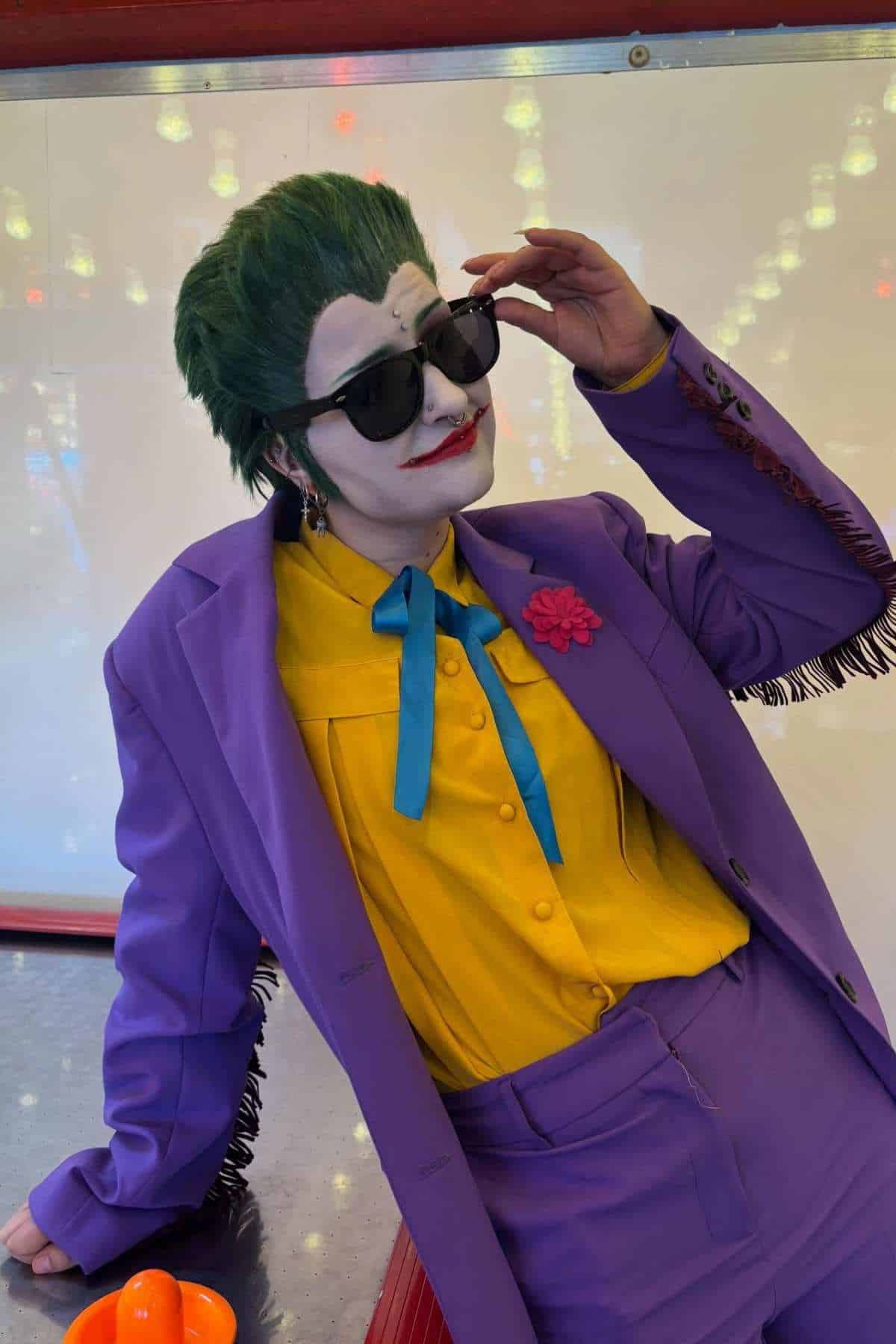 Person in a purple suit, yellow shirt, green wig, and white face paint with red lips, resembling the Joker, poses with dark sunglasses in front of a lit-up background.