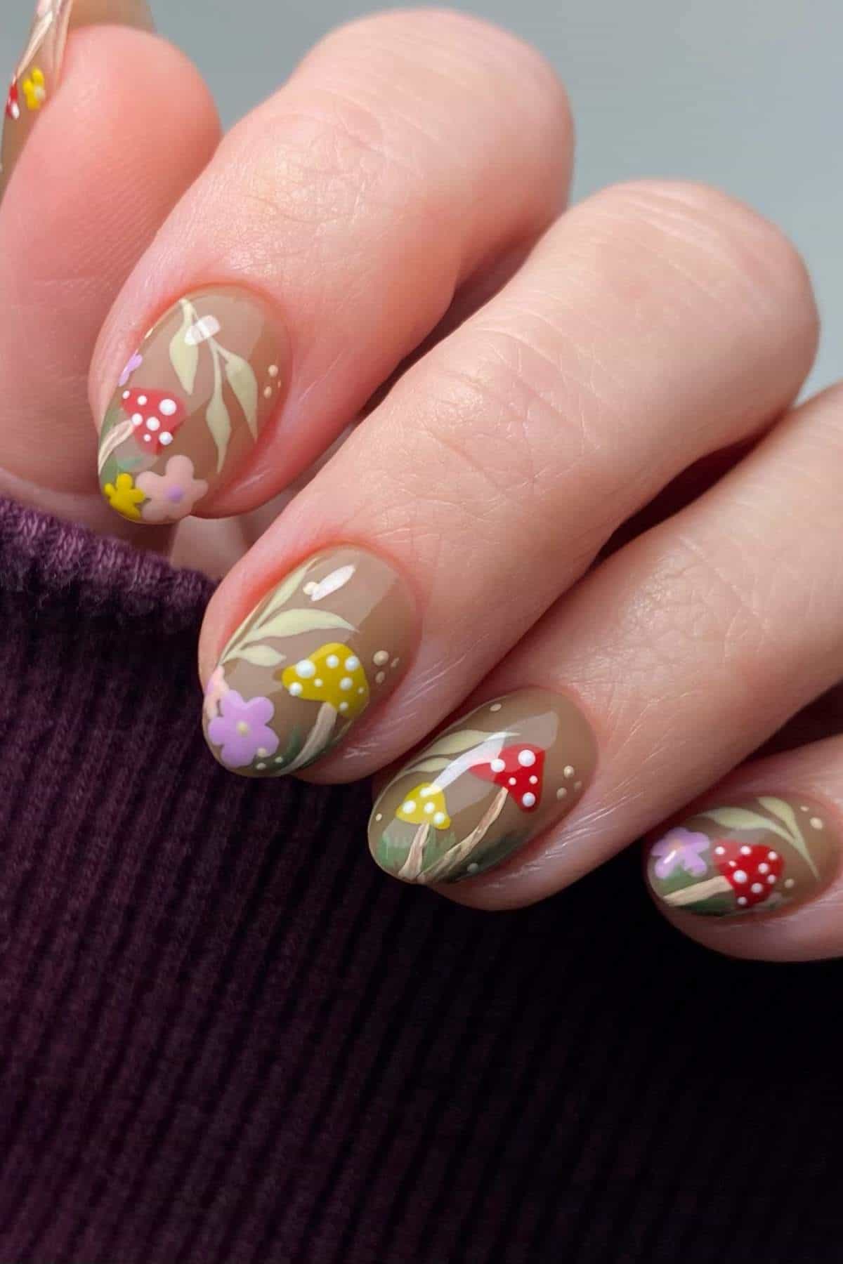 A hand with short nails painted with a clear base and decorated with colorful floral and mushroom designs. The person is wearing a dark purple ribbed sleeve.