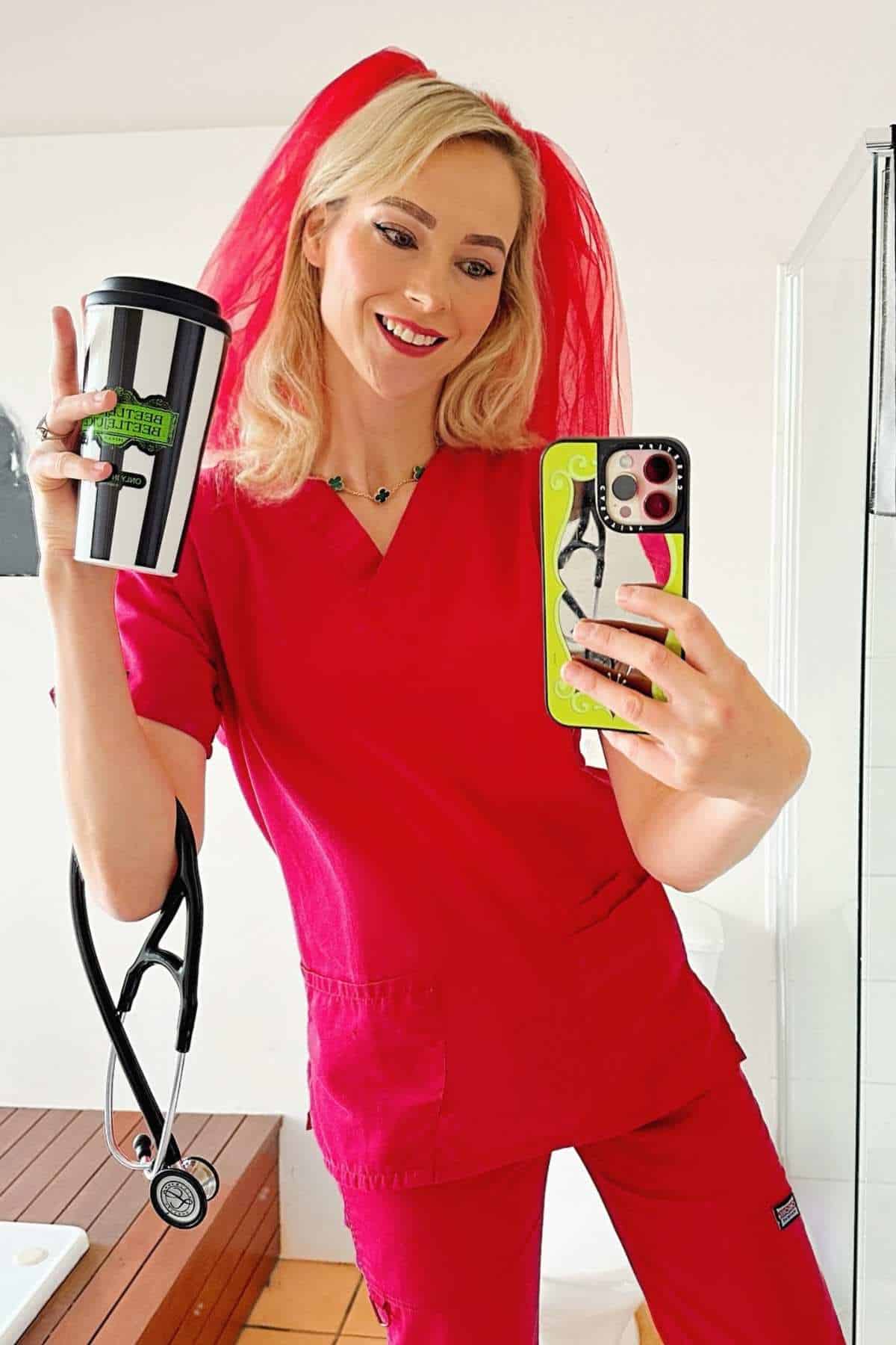 Woman in red scrubs with a stethoscope, holding a striped coffee cup and taking a mirror selfie; she is wearing a red veil and smiling.
