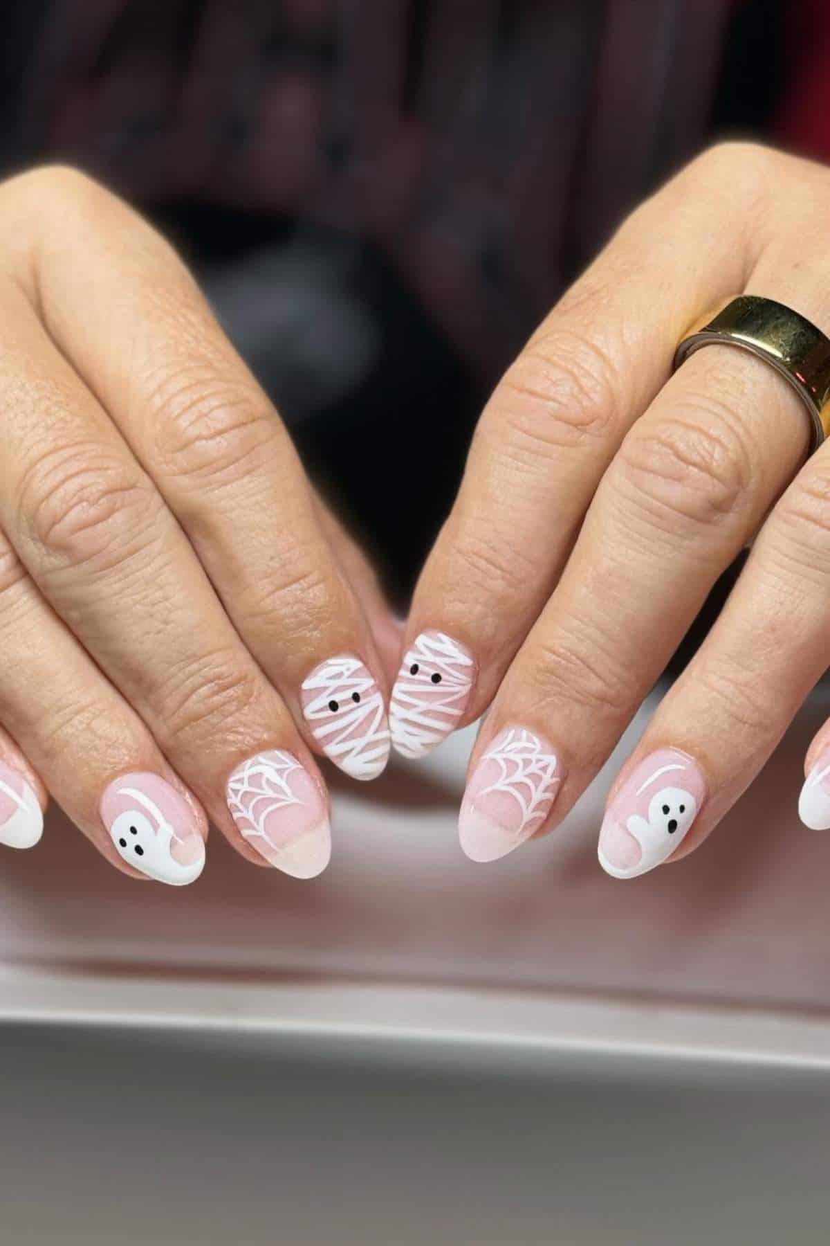Hands with almond-shaped nails featuring Halloween-themed nail art, including ghosts, webs, and mummy designs in pink and white tones.