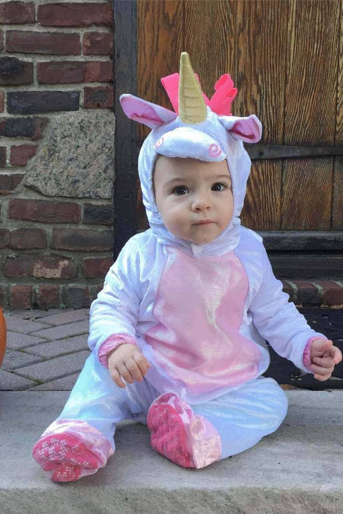 A baby sits on a stone step wearing a unicorn costume with a horn, ears, and pink accents; a wooden door and brick wall are in the background.