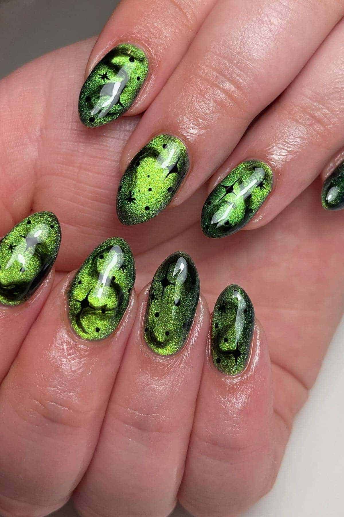 Close-up of hands with oval-shaped nails painted in a shiny green and black abstract design resembling a galaxy or science fiction theme.