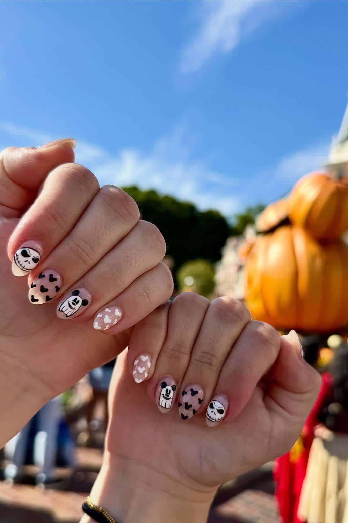 Two hands with Halloween-themed nail art featuring skulls, ghosts, and heart patterns. A large pumpkin decoration and trees are visible in the blurred background.