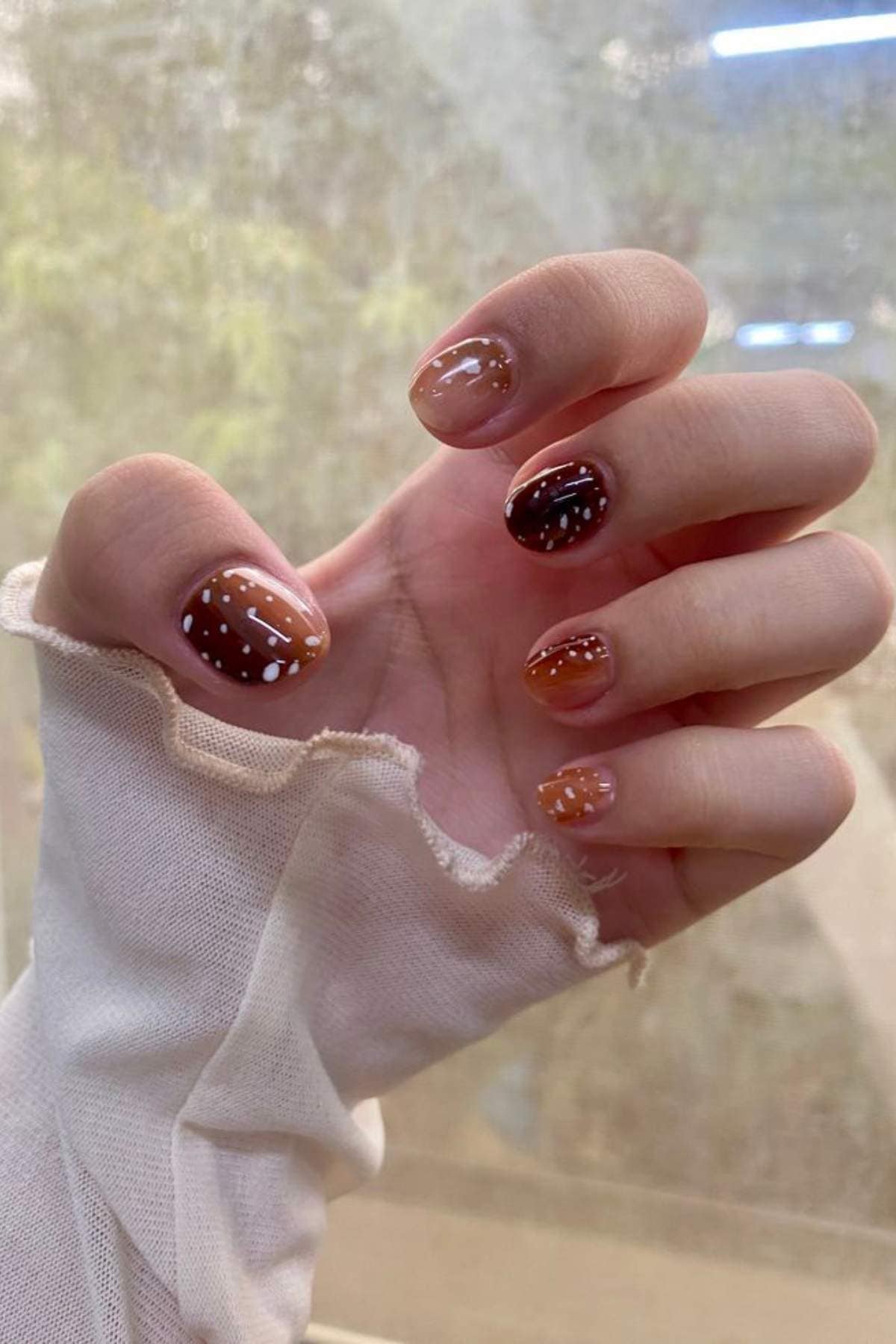 Hand with brown and orange gradient nail polish, white polka dots on each nail, and long-sleeve sheer fabric partially covering the wrist.