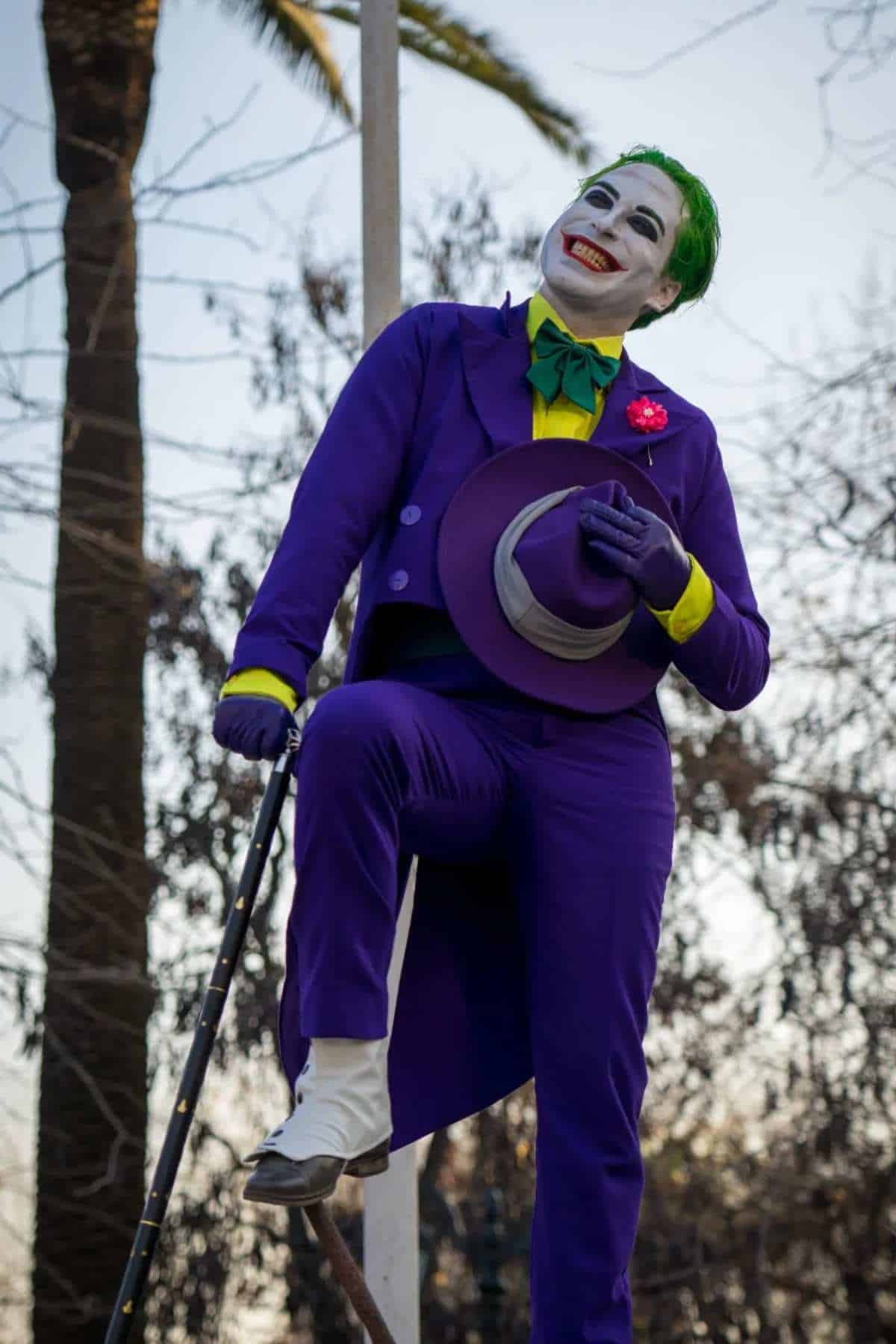 Person in a purple suit, green bow tie, and white gloves poses with a cane and hat, wearing clown makeup with green hair and a wide red smile, outdoors near trees.