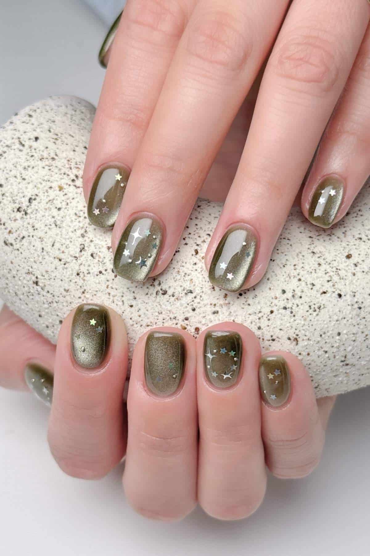 Hands with short, rounded nails painted olive green with a shimmer effect, decorated with small white star-shaped decals, holding a textured white stone.