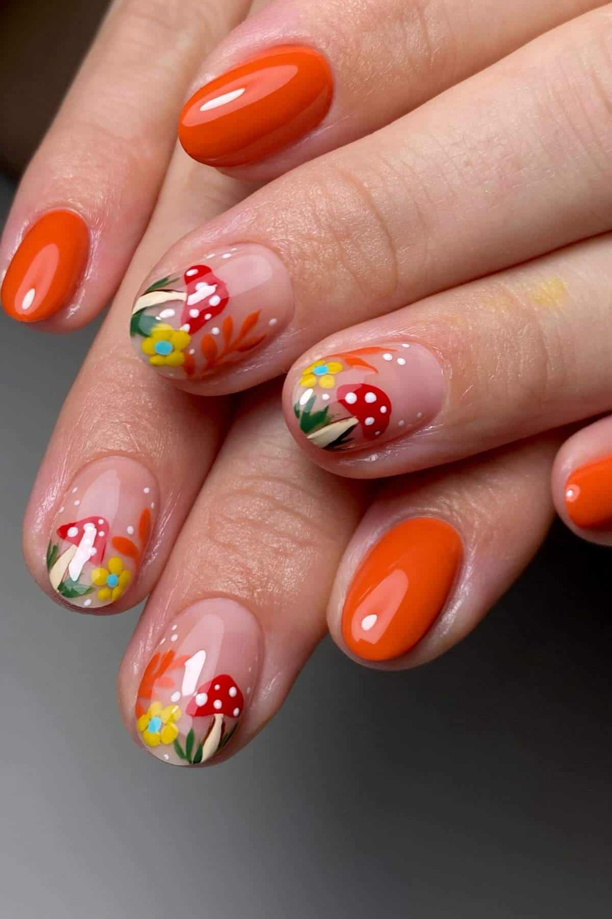 Hand with orange manicured nails, featuring accent nails decorated with red mushrooms, yellow and blue flowers, and green leaves on a clear base.