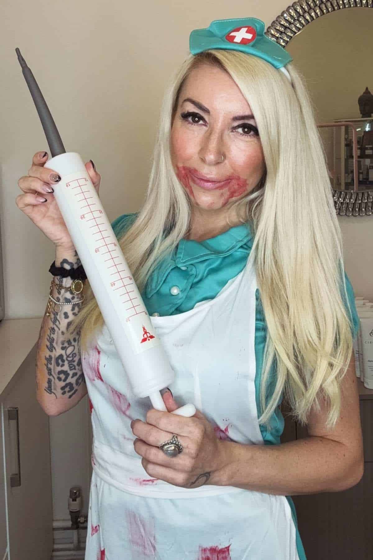 A woman in a nurse costume with smeared makeup holds a large novelty syringe, standing indoors.