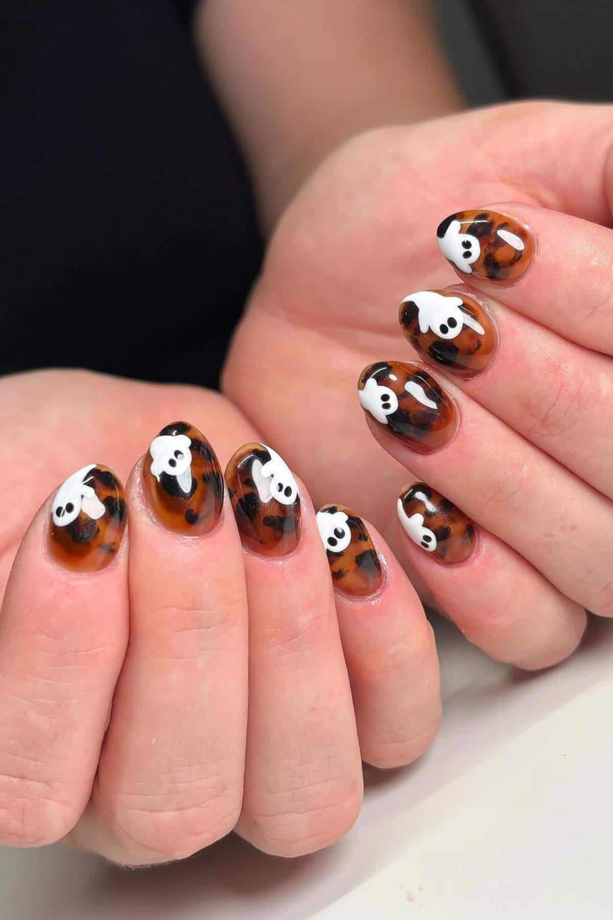 Hands with medium-length nails painted in a brown tortoiseshell pattern, each nail featuring a small white ghost design.