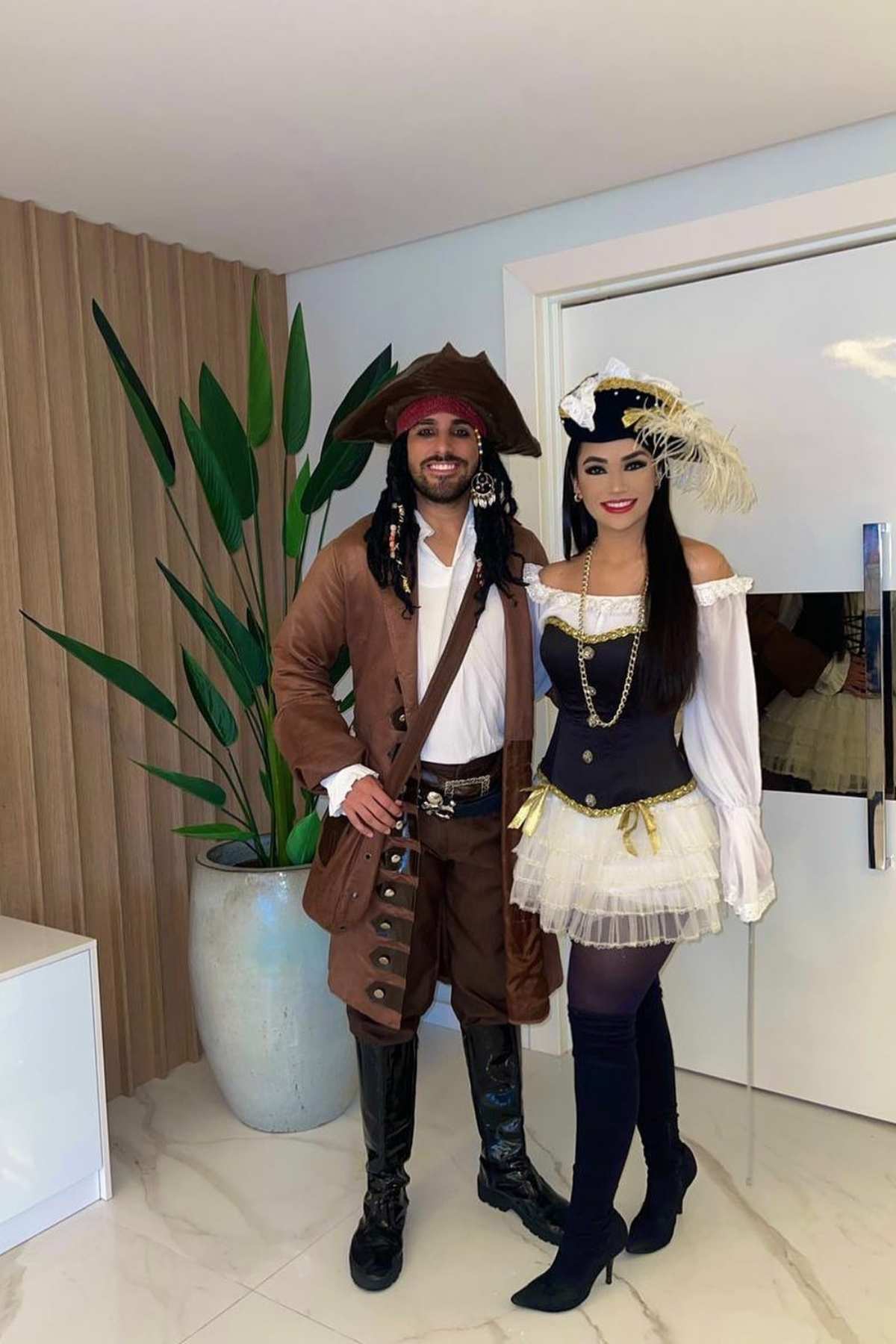 Two people in pirate costumes stand indoors, smiling at the camera; the man wears brown attire and the woman a black and white outfit with a feathered hat. A large potted plant is nearby.