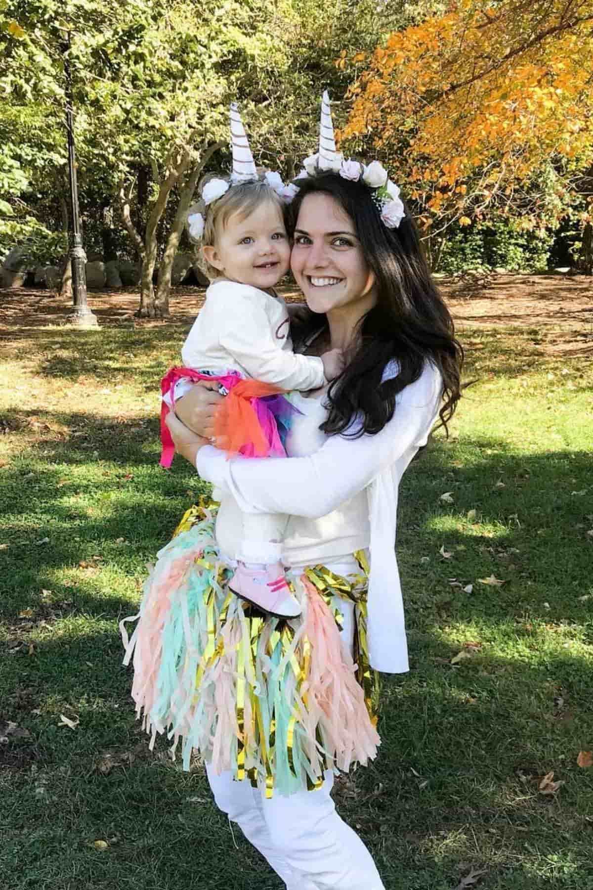 Woman and young girl dressed in unicorn costumes stand outside on grass, smiling at the camera.