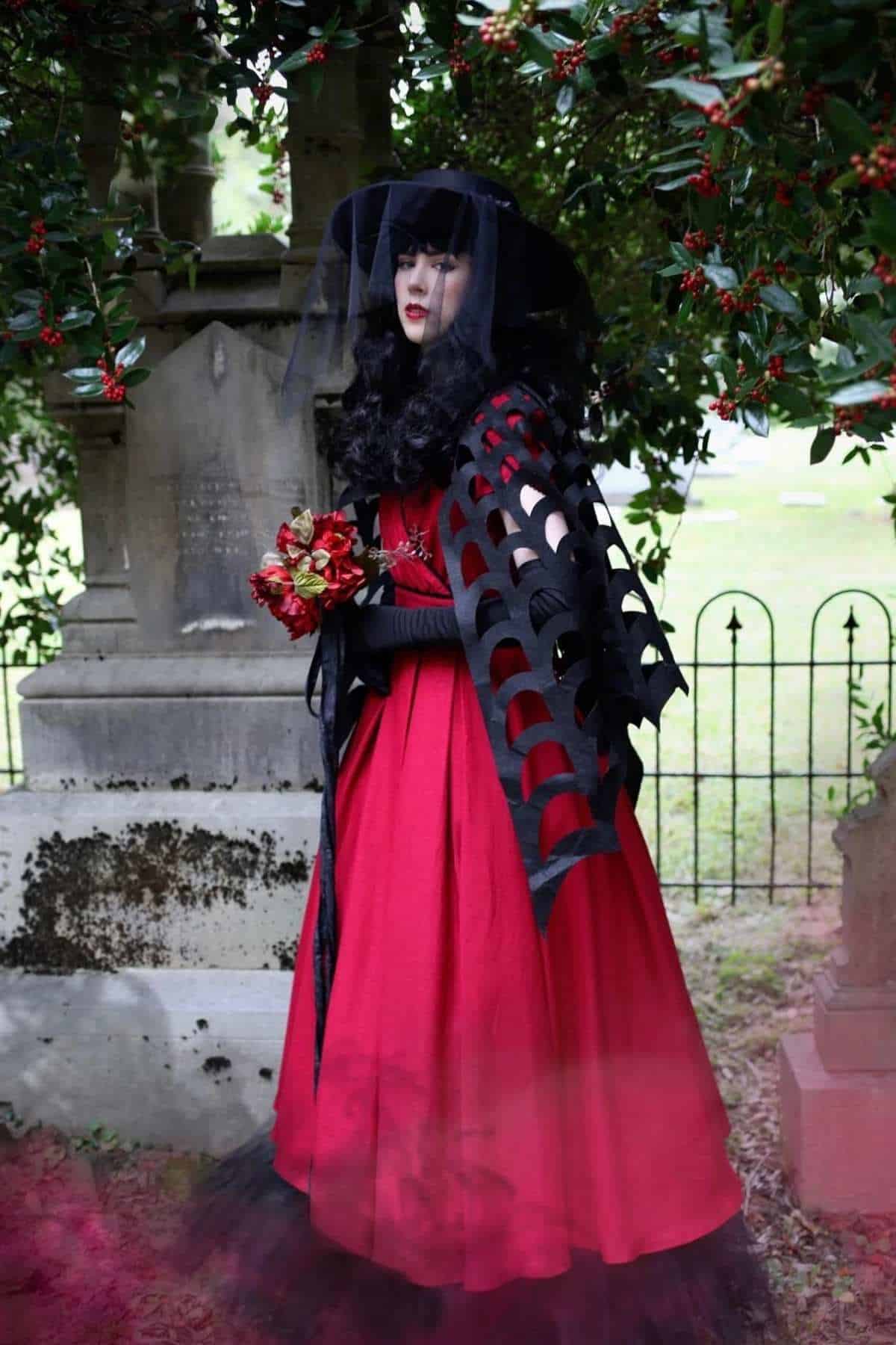 A person in a red dress and black veil stands in a cemetery, holding red flowers, with greenery and gravestones in the background.