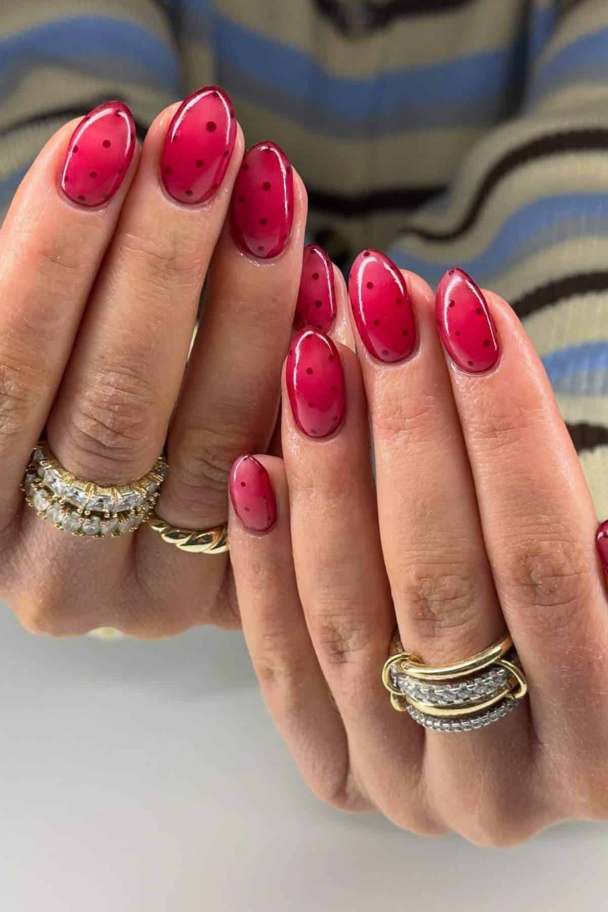 Hands with almond-shaped nails painted pink with black dots, resembling watermelon rind, are shown. The person wears gold and silver rings. Striped sweater sleeves are visible in the background.