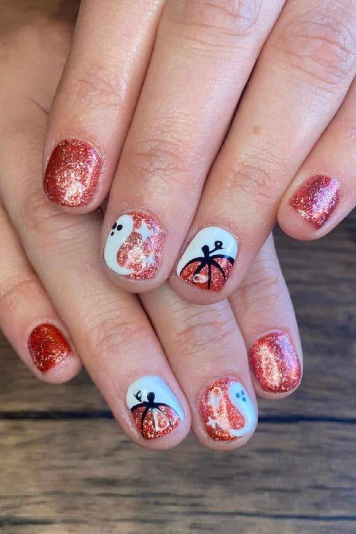 A hand with short, glittery orange nails; some nails feature painted designs of black and white ghosts and pumpkins on a wood surface background.