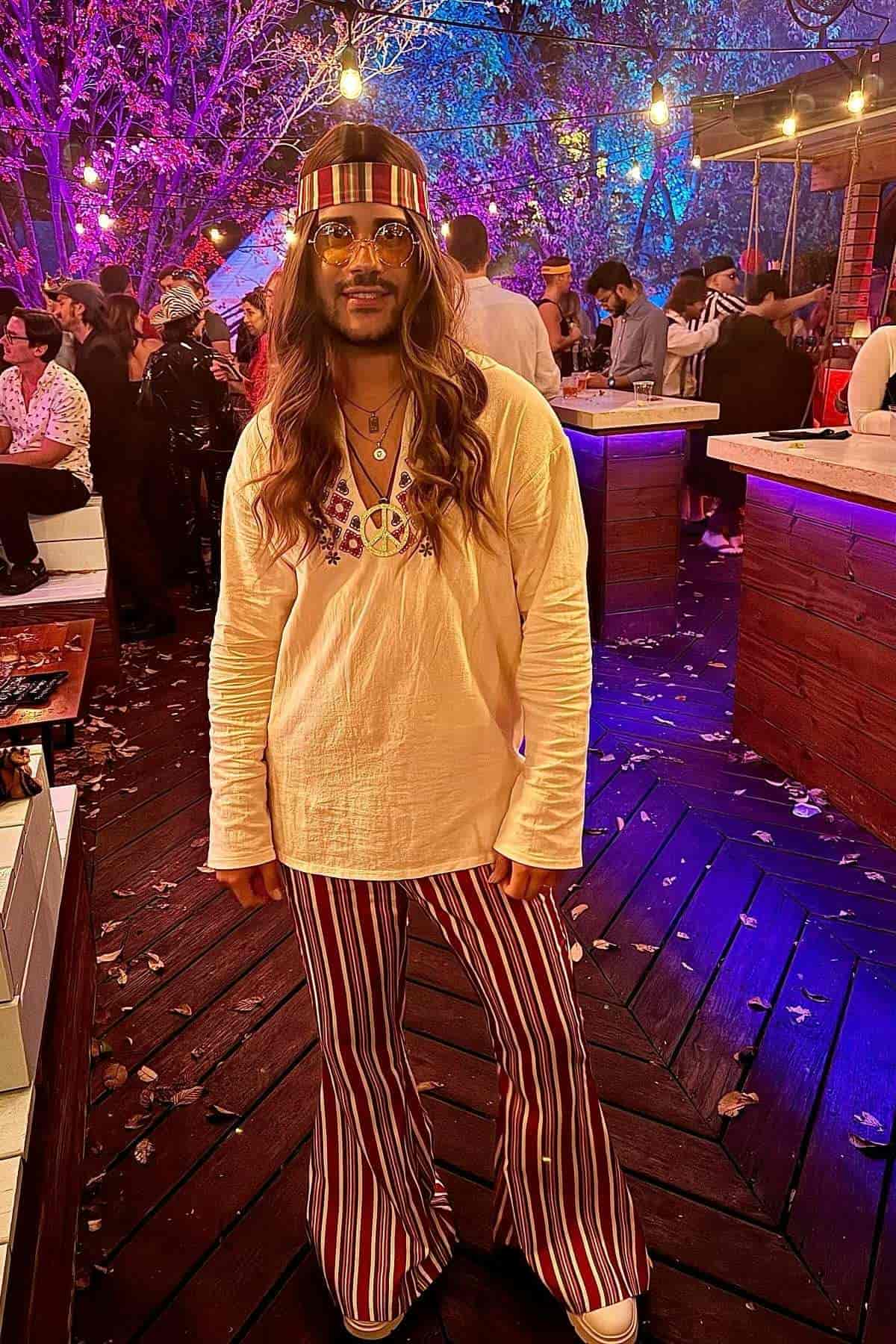 Person stands at an outdoor party in flared striped pants, cream tunic, headband, and peace symbol necklace. Colorful lights and other people are in the background.