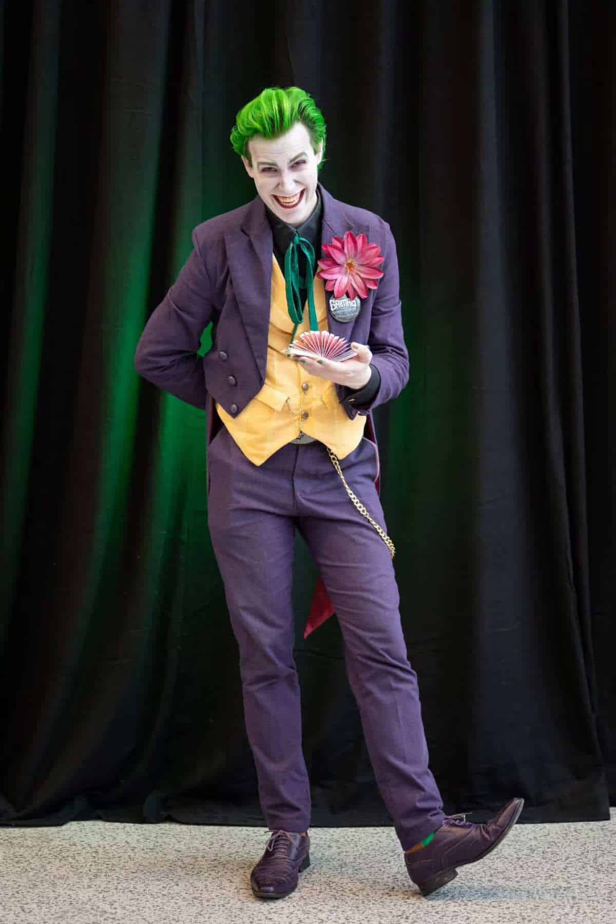 Person dressed as the Joker stands in front of a black curtain, holding a fan of playing cards, wearing a purple suit, yellow vest, green hair, and white face makeup.