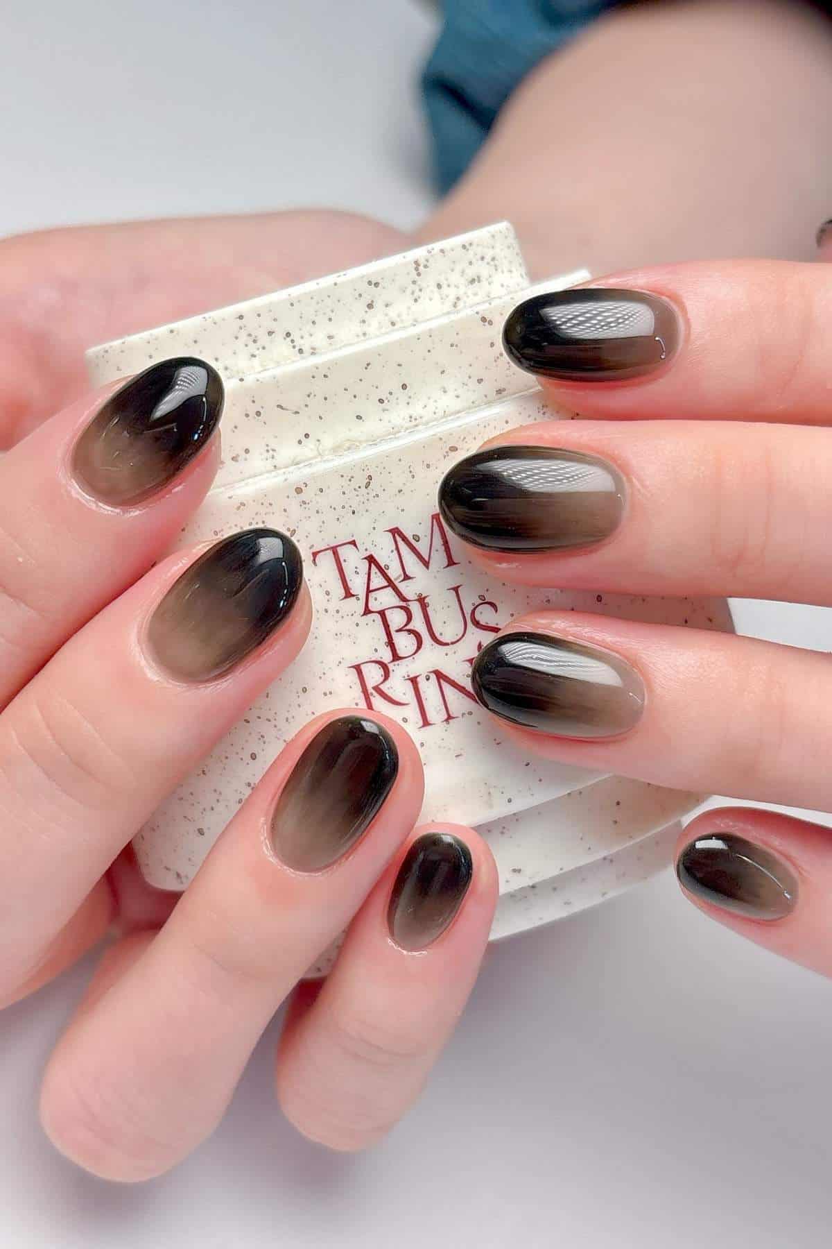 Close-up of hands with oval-shaped nails painted in a glossy black-to-clear ombre design, holding a speckled ceramic cup.
