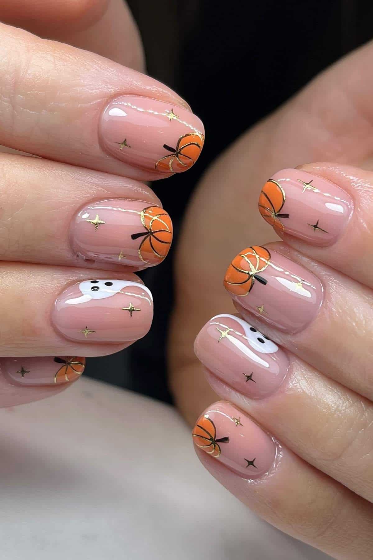Fingernails with nude polish feature painted pumpkins, ghosts, and gold stars as part of a Halloween-themed manicure.