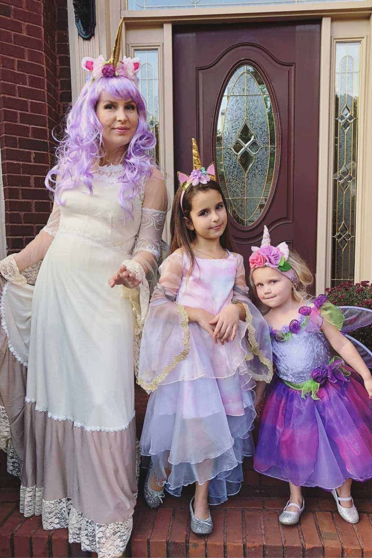 Three people, an adult and two children, stand on a porch dressed in pastel and purple unicorn-themed costumes, posing for a photo in front of a decorative front door.
