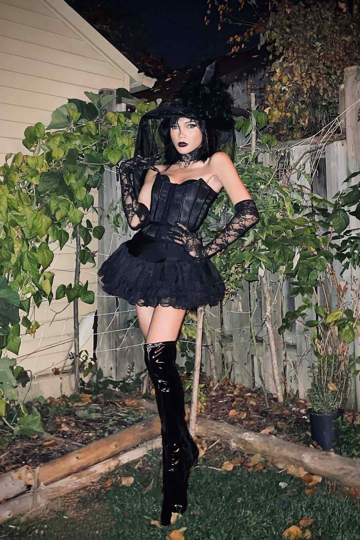Person in a black witch costume with lace gloves and tall boots stands in a backyard near a wooden fence and green plants.