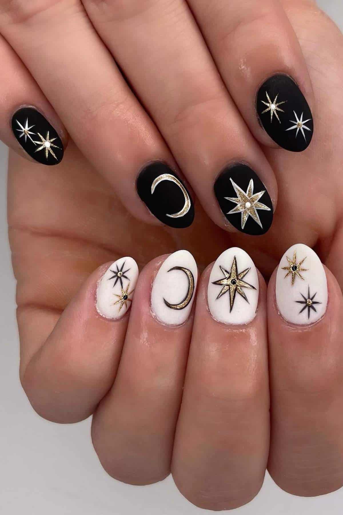 Close-up of hands with manicured nails; black nails feature stars and a crescent moon, white nails feature stars and a crescent moon, all with gold and black celestial designs.