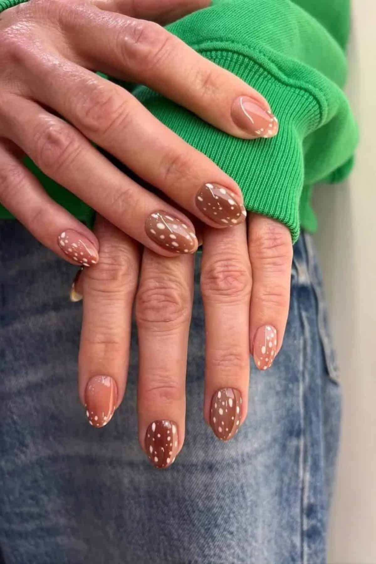 Hands with almond-shaped nails painted nude, decorated with white dotted patterns, are shown resting on denim jeans and a green sweatshirt.