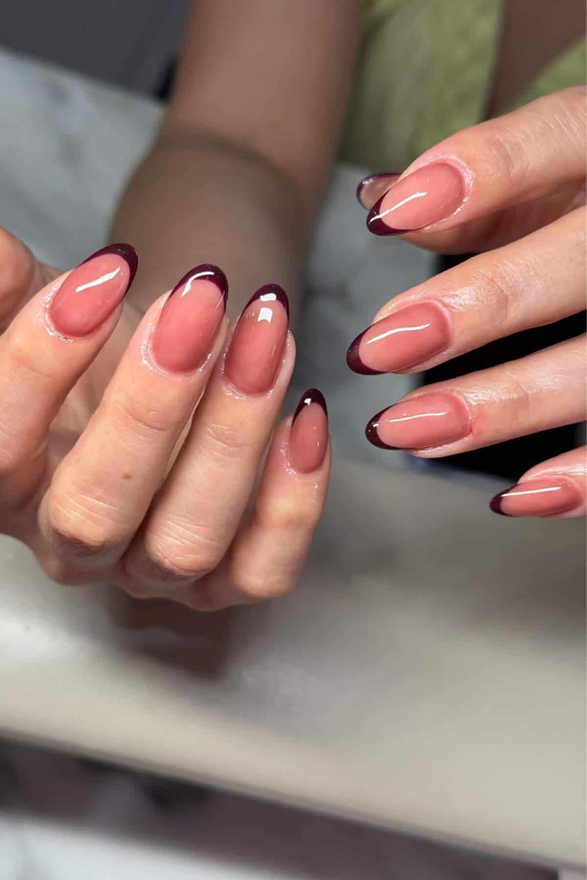 A pair of hands with almond-shaped nails featuring a glossy nude base and dark burgundy French tips.