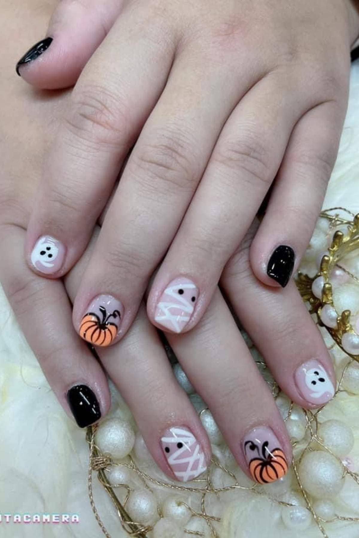Hands with Halloween-themed nail art featuring ghosts, mummies, pumpkins, and black nail polish on a light decorative surface.
