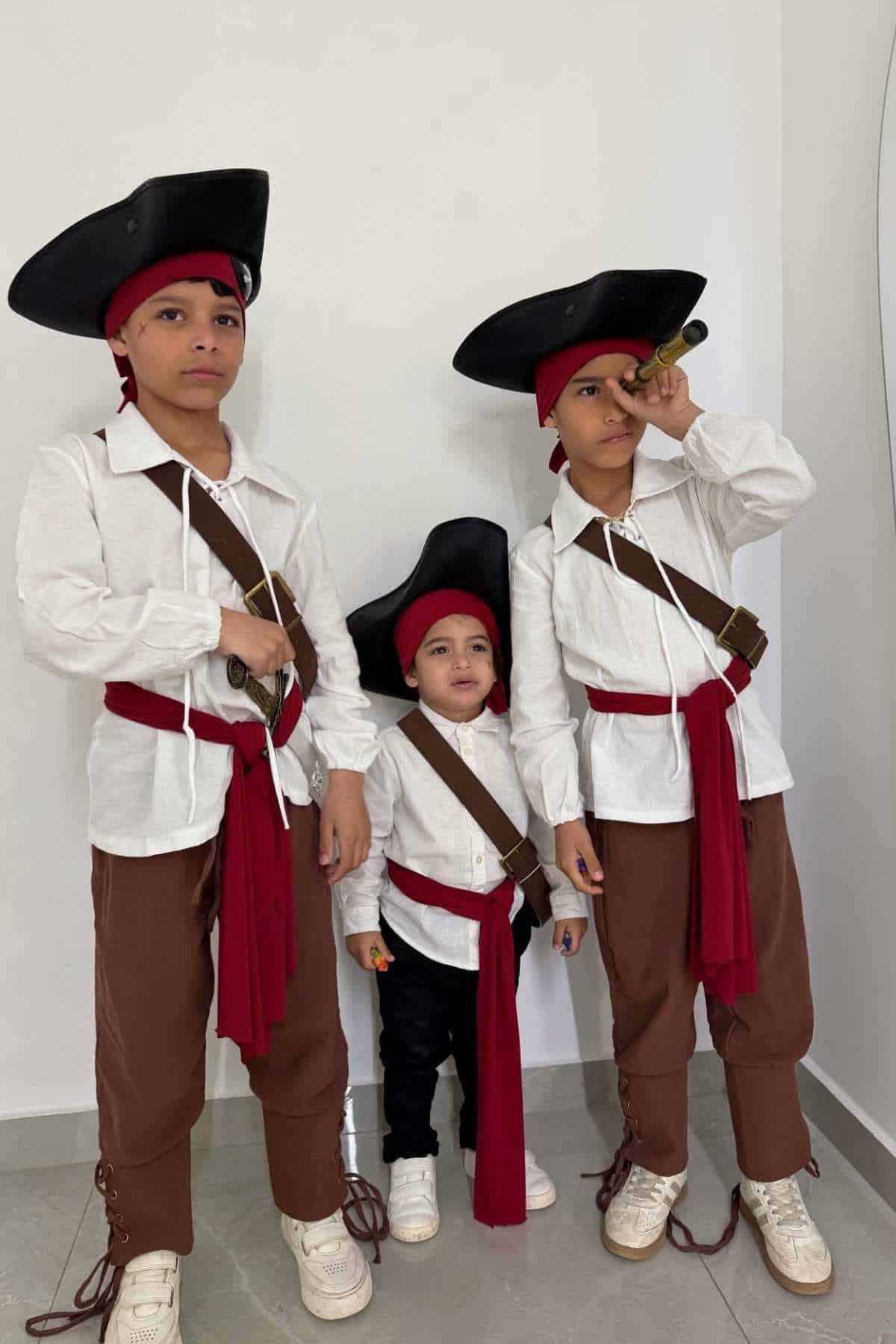 Three children dressed as pirates stand against a white wall; two older kids wear brown pants and one looks through a toy telescope.