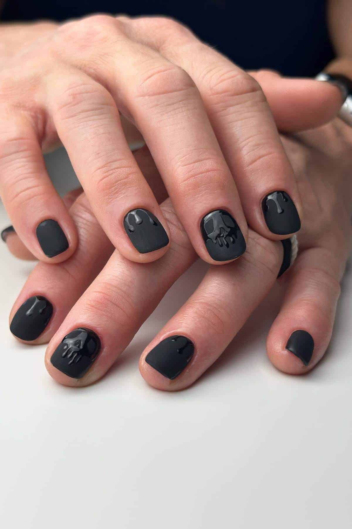 Close-up of hands with short, matte black nails featuring glossy black 3D skull and drip designs on several nails.