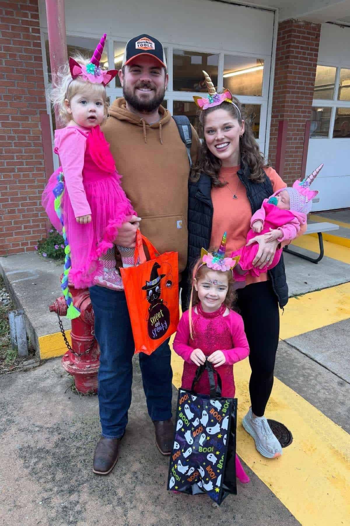 A family with two adults and three young children dressed in unicorn costumes holds Halloween treat bags, standing outside a brick building.