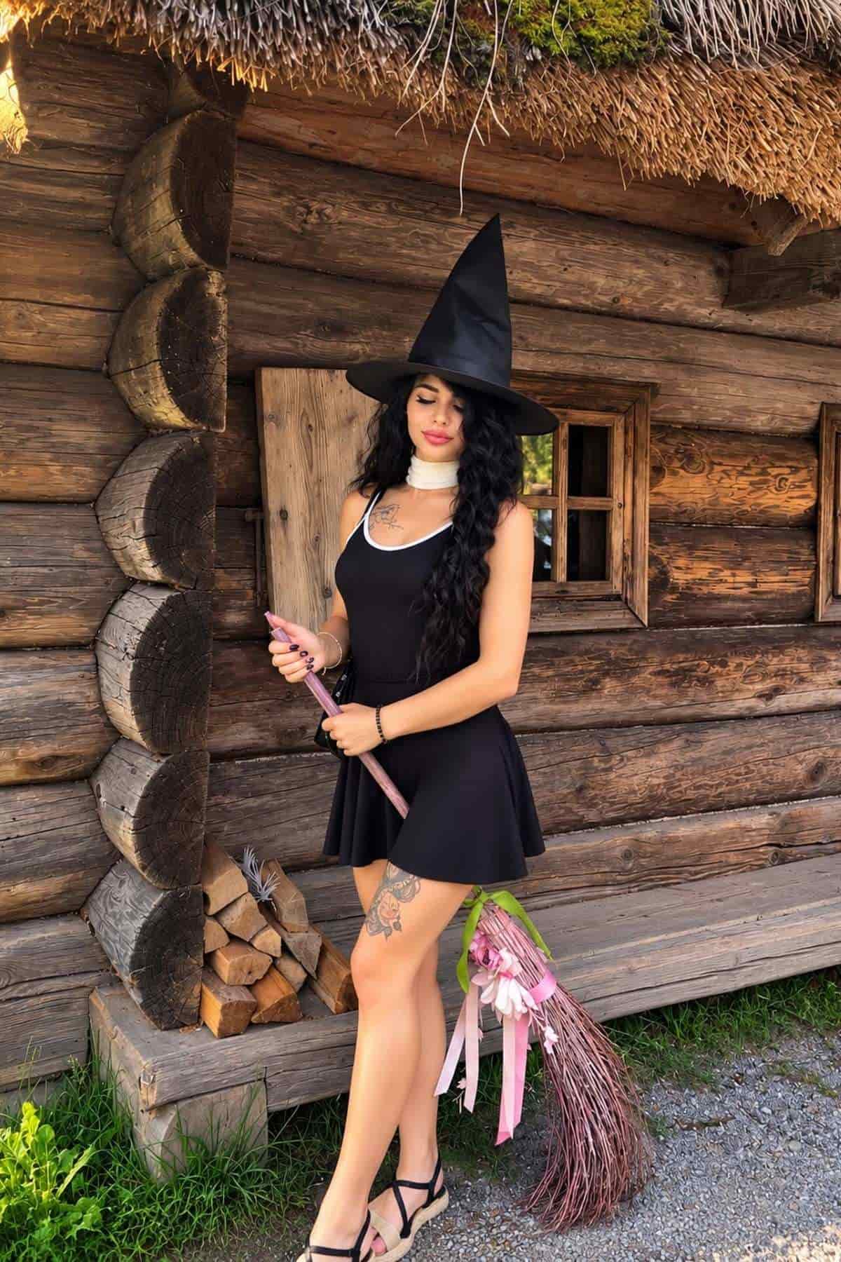 A woman in a black dress and witch hat stands by a wooden cabin, holding a decorated broom with pink ribbons.