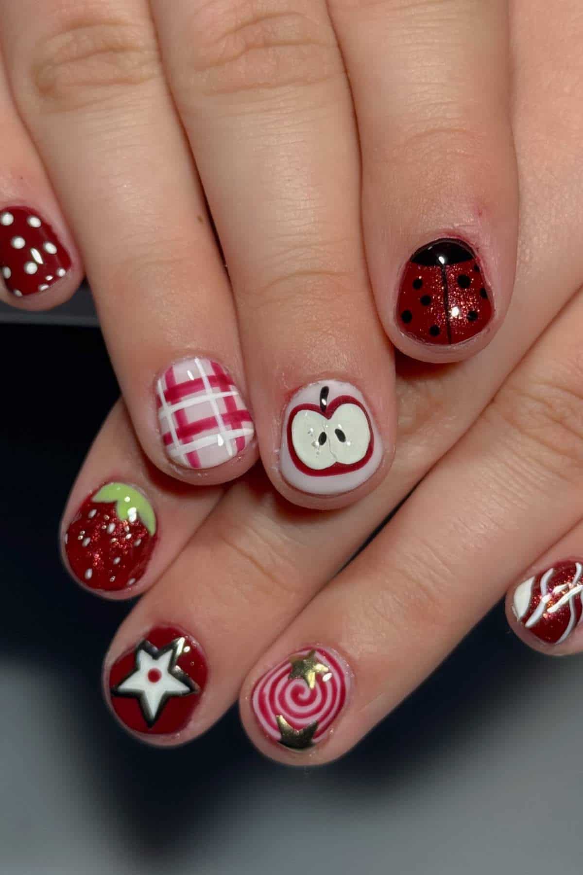 A hand with red and white nail art, featuring designs such as a ladybug, an apple, a star, a swirl, a grid pattern, and polka dots.