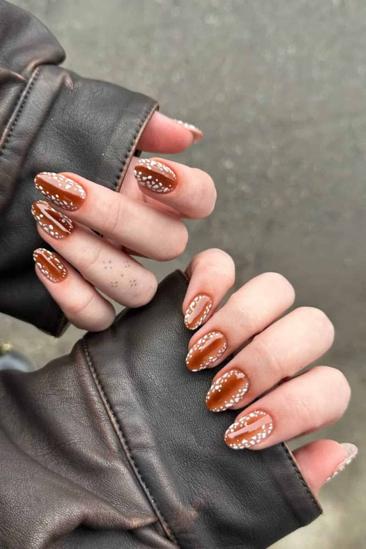 Two hands with almond-shaped brown nails, each outlined with small white dots, are shown emerging from the sleeves of a brown leather jacket.